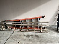 Two ladders side by side against a wall on a concrete floor. One wooden ladder with multiple rungs and one orange aluminum extension ladder.