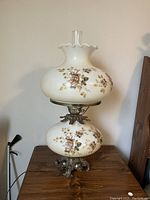 Full view of vintage electric glass lamp with white opaque glass and floral pattern, placed on wooden surface against wall.