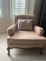 Front angle view of the rose-coloured upholstered occasional chair showing tufted backrest, cylindrical armrest cushions, and wooden legs.