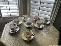 Photo showing 8 assorted fine china teacups each on matching saucers with floral and decorative patterns, arranged on a round table by window light.