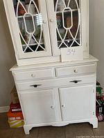 Full front image of the white painted wooden cabinet showing glass doors on the upper section with decorative mullions, two drawers in the middle, and two solid doors on the lower section. The cabinet stands on short sculpted legs with a scalloped base. Shows some wear and paint distress.