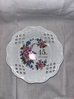 Top view of a white decorative plate featuring floral designs and '45th Anniversary' text with a scalloped edge and lattice cut-out border.