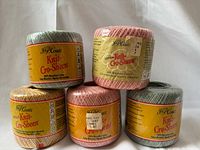 Five spools of J&P Coats Knit Cro-Sheen yarn in almond pink, spruce, and gold colors arranged in a group.