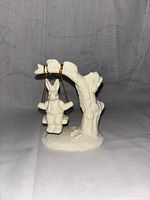 Front view of white ceramic bunny figurine on swing attached to tree branch on base.
