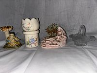 Four decorative items arranged in a row showing detailed colors and design: ceramic cherub candle holder, porcelain floral candle holder set, pink stone-like cherub figurine with dried foliage, and clear glass basket.