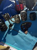 Wide view of all seven skull-themed items arranged on blue surface outdoors, showing mugs, goblets, and a flat skull plaque