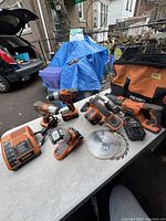 Photo displaying the Ridgid cordless power tools arrayed on a table: reciprocating saw, circular saw with blades, work light, two drills, charger, and batteries, plus a Ridgid carrying case.