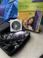 Photo showing Andis EasyClip Groom pet clipper in purple and silver color, power cord, blade guards, user manual DVD, and original product box with image of woman grooming dog.