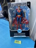 Front view of DC Multiverse Superman action figure in original packaging, including figure and accessories visible through clear plastic window.