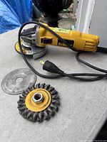 Yellow and black DeWalt corded angle grinder with grinding disk attached, showing side profile and cord plug. Includes wire brush attachments lying on surface.