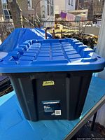 Front view of the black plastic tote with blue lid outdoors showing the size and lid design.
