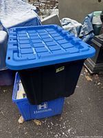 Blue lidded black plastic storage tote photographed outside on top of another blue tote. The lid is securely attached, appears sturdy and in good condition with no visible cracks or major damage.