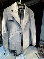 Light gray men's winter coat/jacket hanging showing front side with collar, flap pockets, and side pockets.