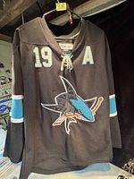 Front view of black San Jose Sharks hockey jersey with #19 and letter A, hanging on a hanger.