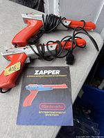 Pair of vintage Nintendo Entertainment System Zapper guns with cables coiled and original instruction manual placed beneath them on a gray surface.