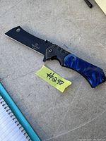 Image of the tactical sharp folding cleaver knife with blade extended, resting on grey surface beside a notebook and yellow lot tag #298.