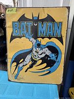 Photo showing a rectangular vintage Batman tin wall art with significant wear and paint chipping on the edges and surface. Batman is depicted in blue and gray costume flying against a yellow background with large Batman text.