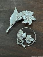 Two sterling silver floral brooches on a dark wooden surface, showing front side details.