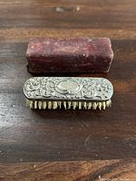 Small antique silver brush with floral and scroll designs on handle, showing intricate craftsmanship and natural bristles underneath.