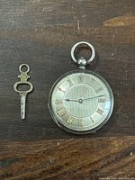 Front view of pocket watch showing silver case, gold Roman numerals on dial, and blue steel hands, alongside the winding key.