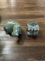 Two carved Asian stone figures displayed on a wooden surface: a greenish stone hippopotamus and a detailed rectangular carved figure with ornamental patterns.