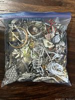 Clear plastic zip-lock bag filled with assorted bracelets, earrings, necklaces, and other jewelry items on wooden surface.