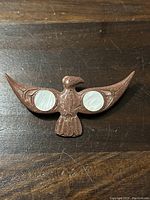 Front view of carved stone bird brooch with white circular shell inlays on wings, laid on wooden surface.