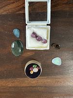 All loose gemstones arranged on wooden surface including star rubies, opals, green cabochon, smoky quartz, and container of various colored stones.