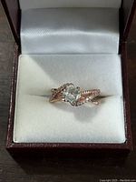 Front view of a rose gold-tone sterling silver ring with a clear cushion-cut center stone surrounded by small clear accent stones on the twisted band, displayed in a white cushioned brown jewelry box.