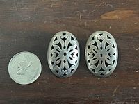 Pair of oval sterling silver clip-on earrings next to a Canadian quarter for size reference, showing cut-out floral design.