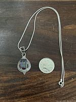 Full view of pendant with chain and quarter for scale, showing detailed silver setting and rectangular mystic topaz stone.