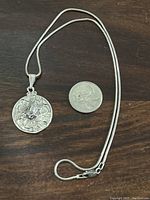 Full necklace with circular embossed pendant next to a coin for size comparison on a wooden surface.