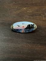 Brooch resting on wooden surface showing front oval painted design of sunset ocean with birds and cliffs.