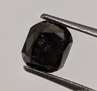 Close-up front view of genuine cushion cut black diamond held by tweezers showing surface facets and dark color.