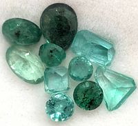 Top-down view of ten faceted genuine emerald gemstones in various cuts and green shades showing natural inclusions.