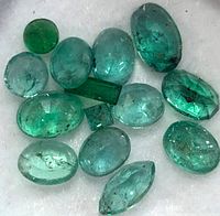Close-up image showing 15 loose emerald gemstones in various shapes including oval, round, marquise, rectangular, and square, displaying a range of green hues with natural inclusions.
