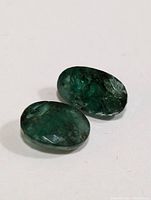 Two oval emerald gemstones lying on a white surface, showing their natural green color and internal inclusions.