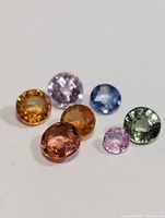 Seven loose faceted fancy color sapphire gemstones in different colors arranged closely on a white background.