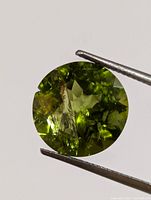 Close-up of round-cut peridot gemstone held with tweezers, showing its olive-green color and internal inclusions.