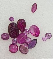 Loose genuine ruby gemstones in multiple shapes and colors arranged on a white surface.