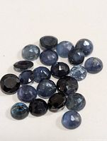 Close-up image showing 24 faceted loose blue sapphires with varied round and oval cuts in shades of light to dark blue.