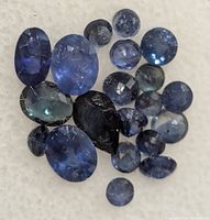 Close-up image showing a mix of various loose, faceted genuine sapphires with blue and bluish-green tones and visible inclusions.