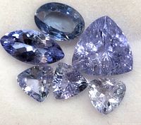 Close-up photo of six loose tanzanite gemstones in various cuts and sizes, showing light to medium blue-purple colors and faceting details.
