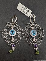 Pair of sterling silver earrings with intricate filigree design, central oval blue topaz stones, small clear stones bordering topaz, round amethyst stones below, and green pear-shaped peridot drop stones. Earrings have hinged hoop closures with clear stones.