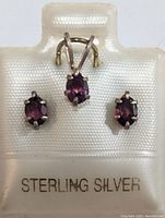 Front view of the garnet pendant and earrings displayed on a white backing marked 'STERLING SILVER'. Shows the oval deep red garnet stones set in silver prongs.