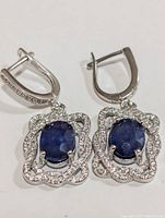 Pair of silver earrings each with oval sapphire center stone surrounded by cubic zirconia in a wavy floral pattern, lever back closure visible.