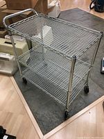 Full view of chrome 3-tier utility cart showing side with handle and wheels on wood and tile floor.