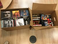 Two cardboard boxes filled with assorted CDs and DVDs placed on a wood floor, showing the general quantity and type of items in the lot.