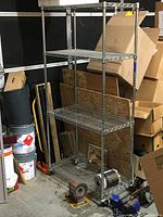 Front view of the 4-shelf chrome metal shelving unit with locking wheels, surrounded by workshop materials and boxes.