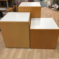 Three square display stands of varying heights, with white tops and wood sides, positioned on a light wood floor in a retail or workshop environment.
